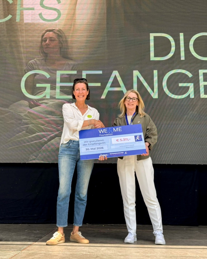 Zwei Frauen stehen lächelnd auf der Bühne und halten einen großen symbolischen Scheck über 5.311 € in der Hand. Im Hintergrund ist ein Bildschirm mit dem Bild einer sitzenden Frau und einem teilweise sichtbaren deutschen Text zu sehen.