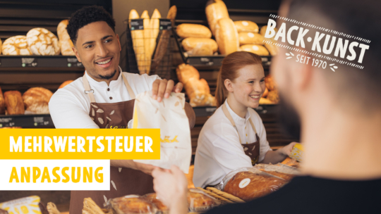 Ein lächelnder Bäckereimitarbeiter reicht einem Kunden eine Papiertüte, während ein weiterer Mitarbeiter neben ihm steht. Im Hintergrund sind Brote in Regalen ausgestellt. Der Text lautet: „MEHRWERTSTEUER ANPASSUNG“, „Mehrwertsteuersenkung“ und „BACK KUNST SEIT 1970“.