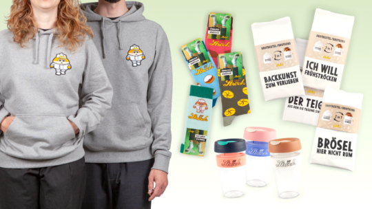 Zwei Personen tragen graue Kapuzenpullis mit einem Bäcker-Cartoon-Motiv. Rechts sind bunte Socken mit Backmotiven, weiße Küchentücher mit deutschen Sprüchen und drei wiederverwendbare Plastikbecher mit pastellfarbenen Deckeln zu sehen.