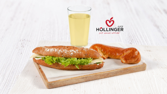 Auf einem Holzbrett liegen ein Sandwich mit Salat und Käse, ein gedrehtes Brötchen und ein Glas Saft. Im Hintergrund ist das Logo von „Höllinger Juice“ zu sehen.