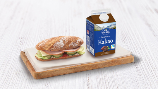 Ein Sandwich mit Schinken, Käse, Gurke und Tomate auf einem kleinen Holzbrettchen neben einem Karton Schokoladenmilch mit der Aufschrift „Kakao“, alles auf einer weißen Unterlage.