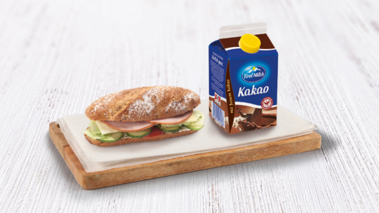 Ein Sandwich mit Schinken, Käse und Gurke auf einem knusprigen Brötchen liegt auf einem Holzbrett neben einem Karton „Tirol Milch Kakao“-Schokoladenmilch.