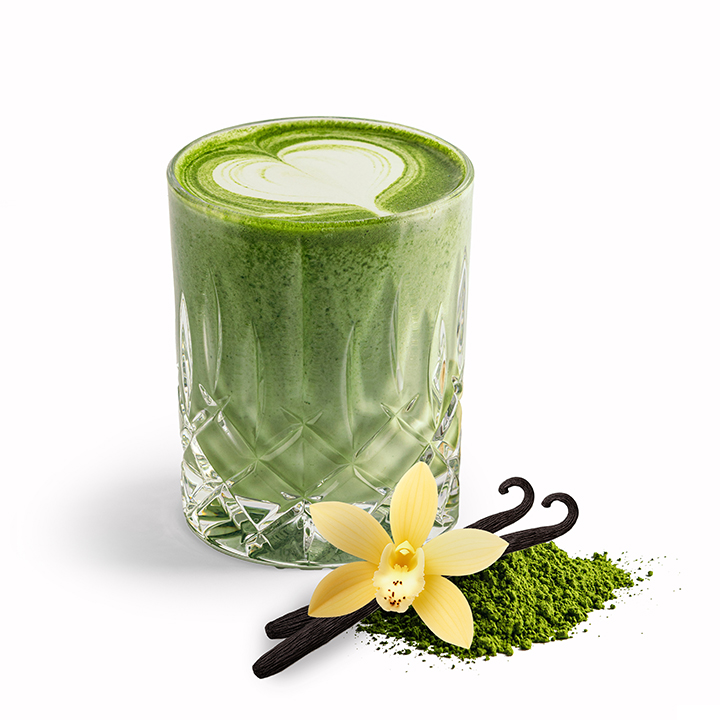 Ein Glas Matcha-Latte mit herzförmiger Schaumverzierung, garniert mit einer Vanilleblüte, zwei Vanilleschoten und einem kleinen Häufchen Matcha-Pulver.