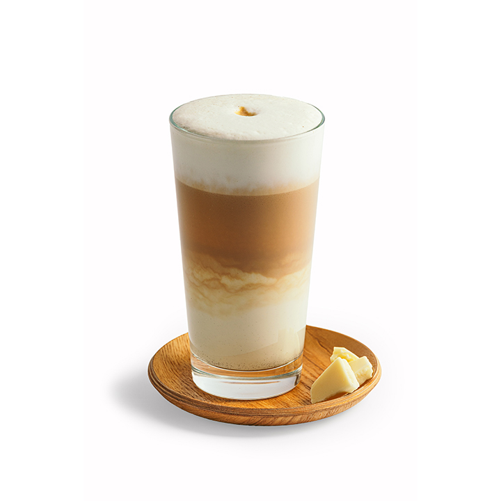 Ein großes Glas geschichteter Kaffee mit Schaum auf einem Holzuntersetzer, dazu zwei Stückchen weiße Schokolade.