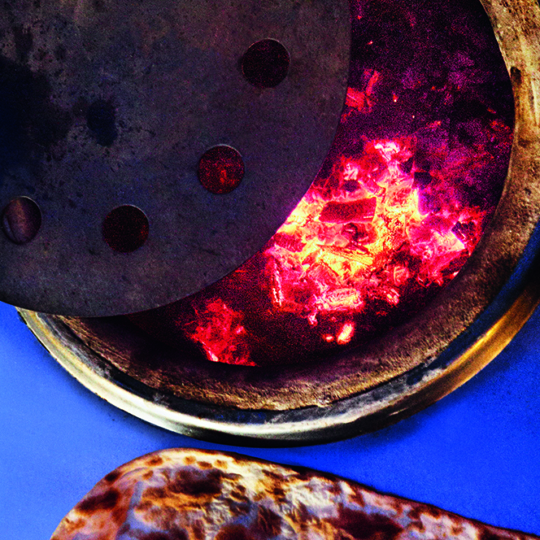 Ein Tandoor-Ofen mit glühenden Kohlen im Inneren ist teilweise mit einem Metalldeckel abgedeckt. Im Vordergrund ruht ein Stück Naan-Brot auf einer blauen Fläche in der Nähe.