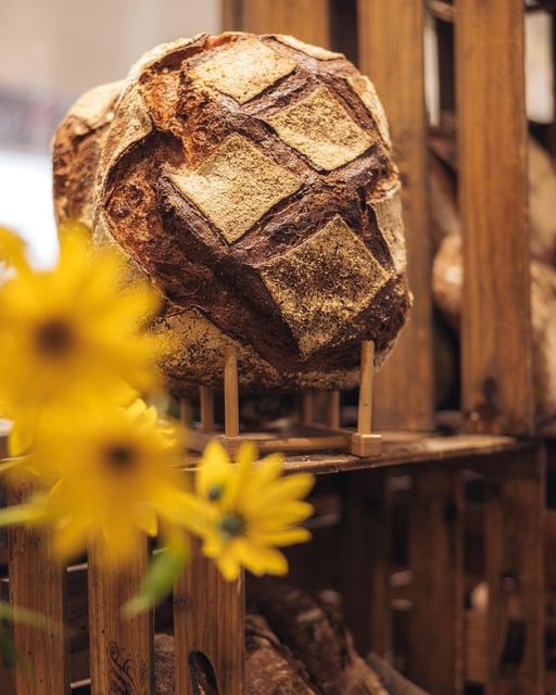 Ein rustikaler Laib handwerklich hergestellten Brotes mit rautenförmiger Kruste wird aufrecht auf einem Holzgestell präsentiert. Leuchtend gelbe Blumen sind im Vordergrund verschwommen.