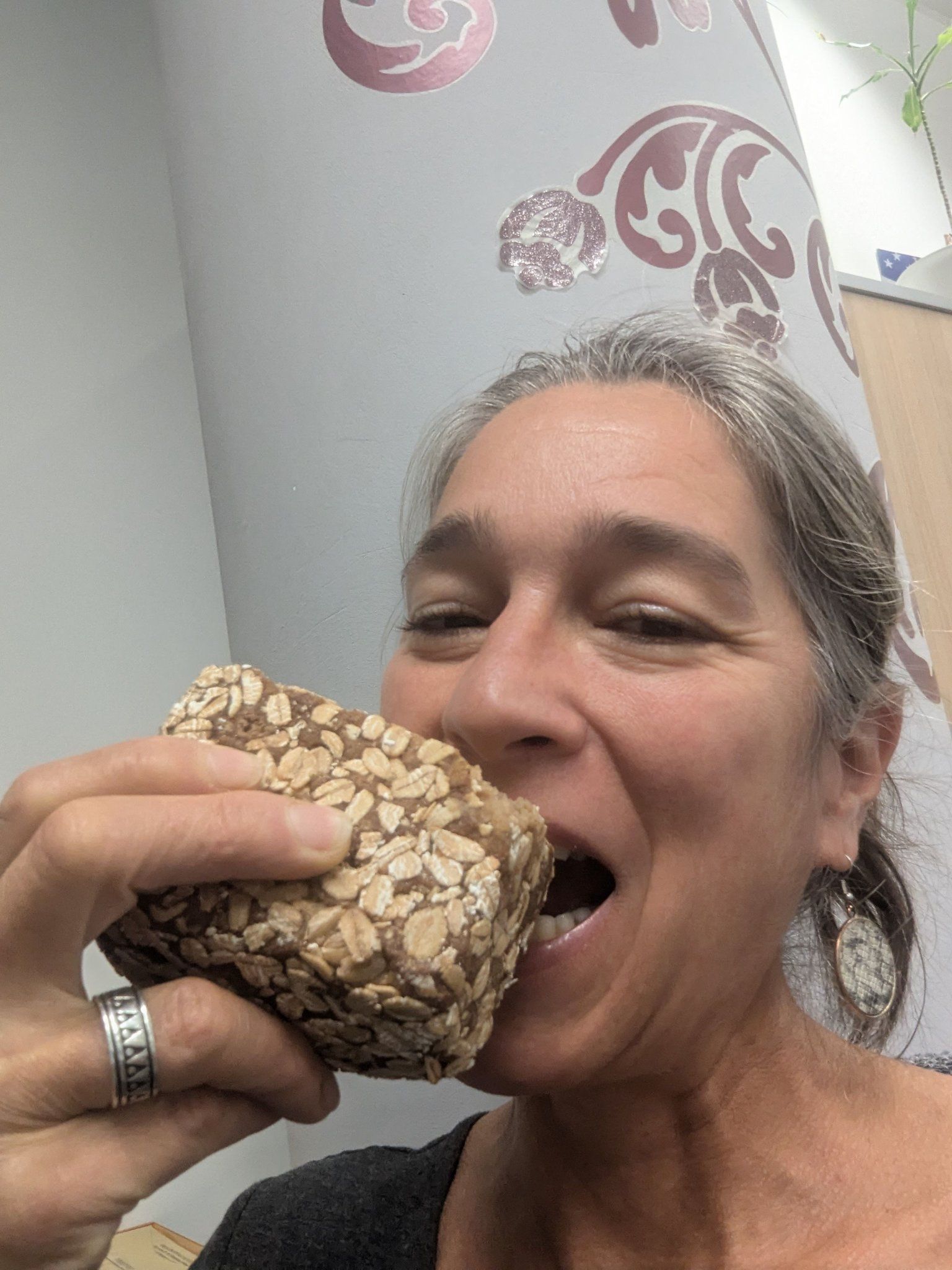 Eine Frau mit grauem Haar hält ein mit Haferflocken belegtes Stück Brot an den Mund, als wolle sie hineinbeißen. Sie steht in einem Innenraum vor einer verzierten Wand.
