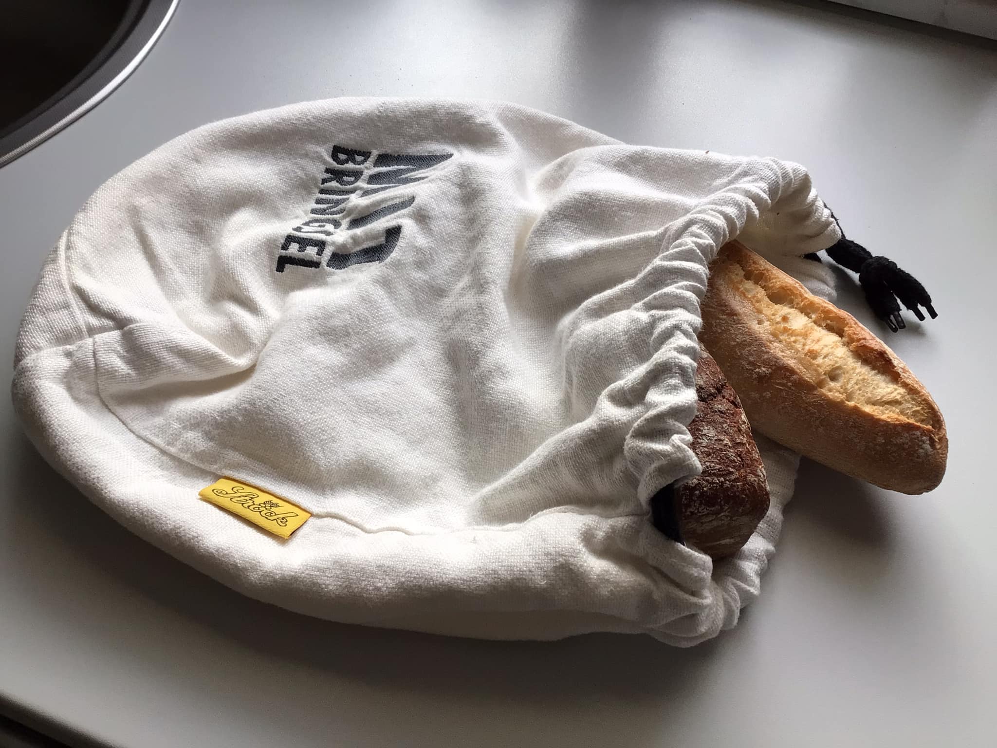 Zwei Baguettes, ein helles und ein dunkles, liegen teilweise in einer weißen Tasche mit Kordelzug auf einer Küchenzeile. Die Tüte ist mit schwarzer Schrift und einem gelben Etikett versehen.
