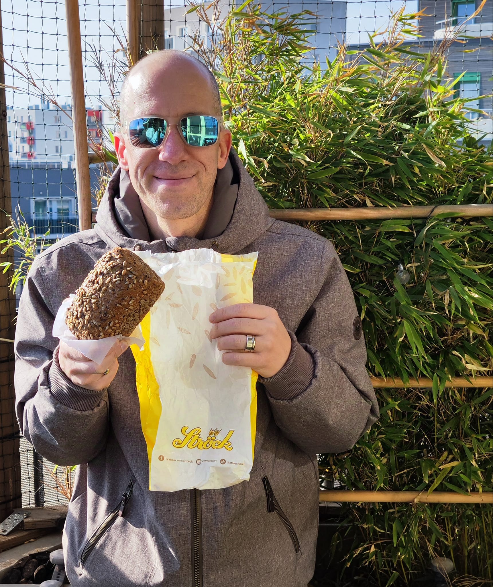 Eine Person mit Sonnenbrille und grauer Jacke lächelt, während sie einen Laib Körnerbrot in einer gelb-weißen Papiertüte hält. Im Hintergrund sind grüne Bambuspflanzen und ein Gebäude zu sehen.