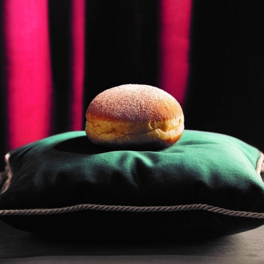 Ein einzelner gepuderter Donut sitzt auf einem plüschigen grünen Samtkissen mit goldenen Quasten vor einem dunklen Hintergrund mit tiefrosa Längsstreifen.