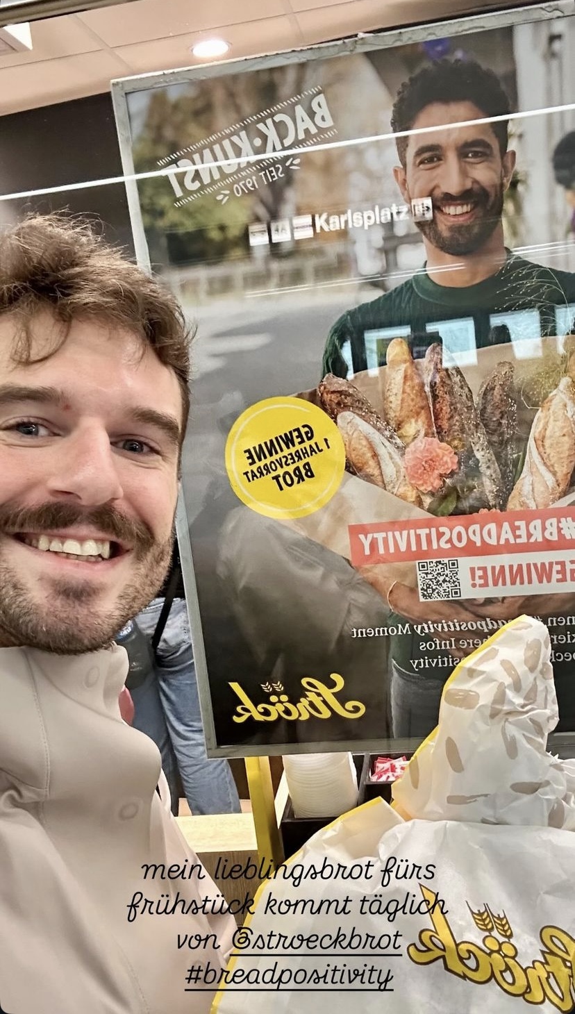 Ein lächelnder Mann macht ein Selfie vor einem Bäckereiplakat, auf dem ein anderer lächelnder Mann mit verschiedenen Brotsorten abgebildet ist. Auf der Theke stehen Tüten mit Backwaren. Der deutsche Text über das Lieblingsfrühstücksbrot ist unten eingeblendet.