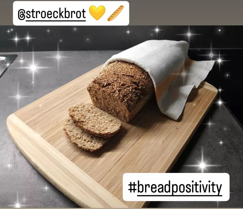 Ein Laib Vollkornbrot, teilweise mit einem weißen Tuch bedeckt, liegt auf einem hölzernen Schneidebrett, von dem zwei Scheiben abgeschnitten sind. Dekorative Glitzer und Textüberlagerungen lesen @stroeckbrot 💛🍞 und #breadpositivity.