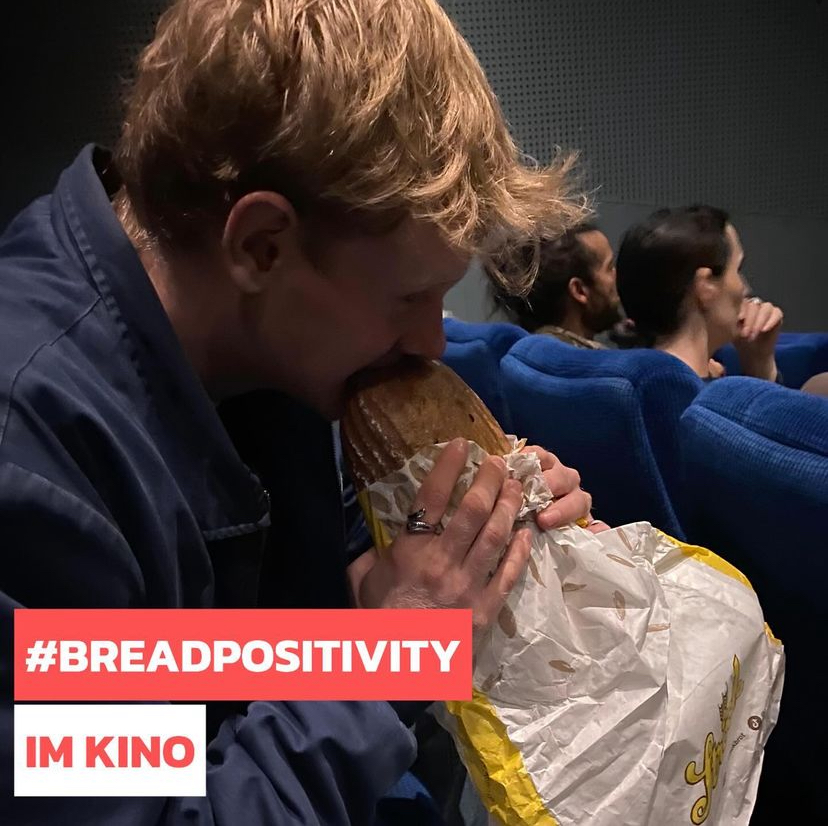 Eine Person mit hellem Haar sitzt in blauen Theatersesseln und beißt begeistert in ein großes, in Papier eingewickeltes Brot. Andere sitzen daneben. Der Text #BREADPOSITIVITY IM KINO ist in fetten roten und weißen Lettern überlagert.