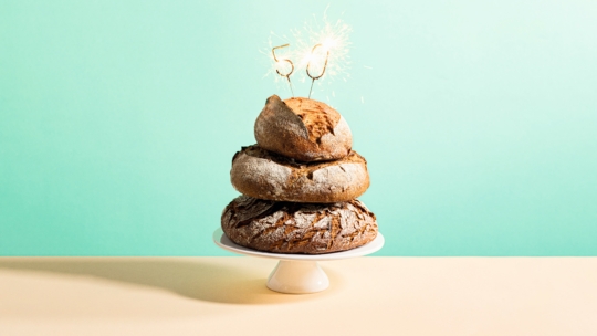Ein Stapel von drei runden Brotlaiben auf einem weißen Kuchenständer, mit brennenden Wunderkerzen in Form der Zahl 50 auf der Spitze, vor einem hellblauen Hintergrund.
