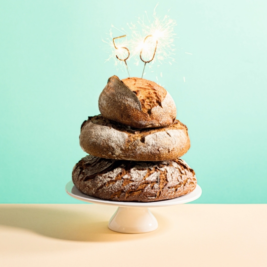 Ein Stapel von drei runden Brotlaiben auf einem weißen Kuchenständer, mit brennenden Wunderkerzen in Form der Zahl 50 auf der Spitze, vor einem hellblauen Hintergrund.