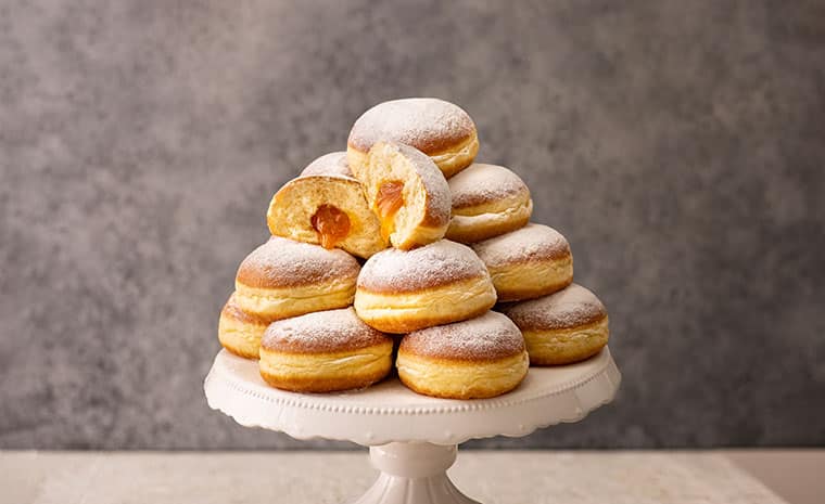 Ein Stapel mit Gelee gefüllter Krapfen ist auf einem weißen Kuchenständer angeordnet; ein Krapfen oben ist aufgeschnitten, um eine Orangenmarmeladenfüllung freizulegen. Der Hintergrund ist ein strukturiertes Grau.