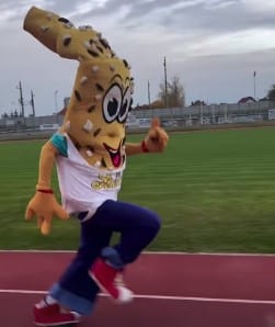 Eine Person in einem riesigen Cartoon-Brot-Maskottchen-Kostüm läuft in blauen Hosen, roten Schuhen und einem weißen Hemd auf einem Sportplatz. Der Hintergrund zeigt Gras, einen Zaun und einen bewölkten Himmel.