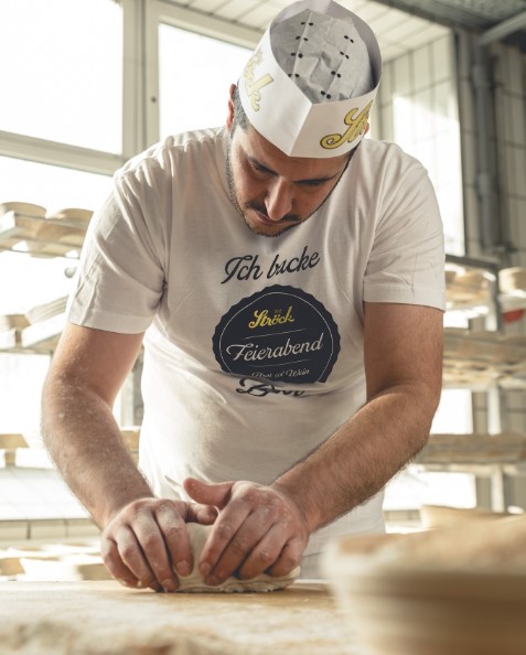 Ein Bäcker, der einen Papierhut und ein weißes T-Shirt mit deutscher Aufschrift trägt, knetet Teig auf einer bemehlten Fläche in einer Bäckerei, mit Regalen voller Brot im Hintergrund.