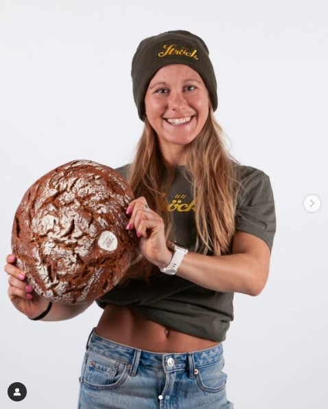 Eine lächelnde Frau mit Mütze und T-Shirt hält einen großen runden Laib Brot. Sie steht vor einem schlichten weißen Hintergrund und trägt eine blaue Jeans und eine Uhr.