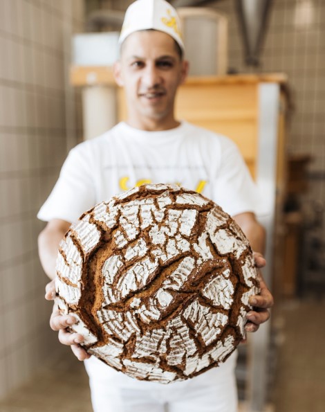 Ein Bäcker in weißer Uniform und mit Hut hält einen großen runden Laib rustikalen, handwerklich hergestellten Brotes mit einer rissigen, mehlbestäubten Kruste vor sich. Im Hintergrund ist eine geflieste Bäckerei zu sehen, die leicht unscharf ist.