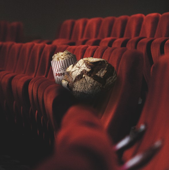 Ein Becher Popcorn und ein Laib rustikales Brot sitzen auf roten Kinosesseln, der Fokus liegt auf dem Essen und die anderen Sitze um sie herum sind leer.