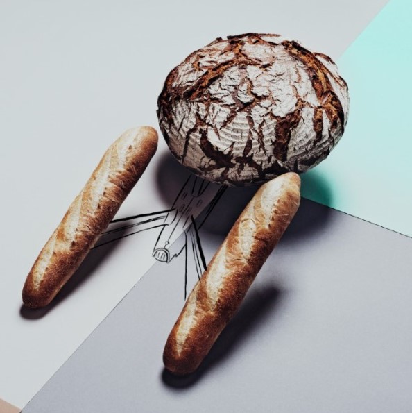 Ein runder Brotlaib und zwei Baguettes sind auf einer Fläche mit minimalistischen Strichzeichnungen angeordnet, so dass die Brote an einen Menschen erinnern, der mit Holzbeinen geht.