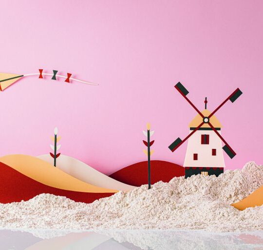 Eine skurrile Szene mit Papierkunsthügeln, einer Windmühle und Blumen auf einer sandigen Fläche vor einem rosa Hintergrund. Ein bunter Papierdrachen fliegt in den Himmel.