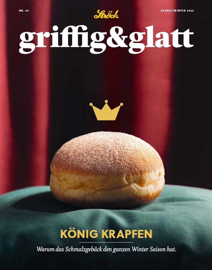 Ein einzelnes gezuckertes Gebäck (Krapfen) sitzt auf einer grünen Fläche unter einer gezeichneten goldenen Krone, mit einem dunkelroten Vorhang im Hintergrund. Oben steht "griffig&glatt" und unten "KÖNIG KRAPFEN.".